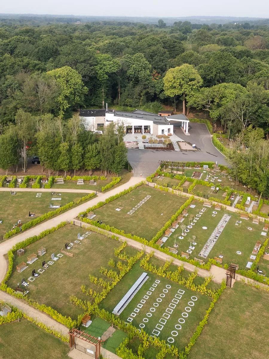 Aerial view of Bluebell Cemetery: rows of grave plots divided by hedged paths, with the white single-storey crematorium building and woodland behind