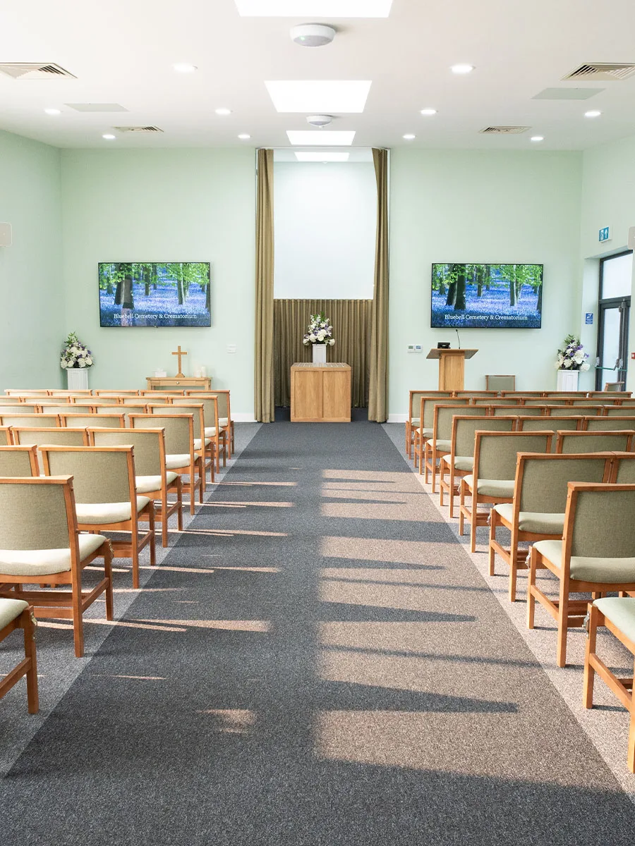 The ceremony room at Bluebell Crematorium: rows of upholstered chairs either side of a central aisle facing a lectern, flowers and two wall screens