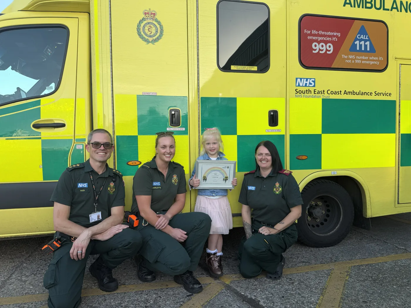 Delilah holding her bravery certificate in front of an ambulance with the SECAmb team who presented it at Paddock Wood Make Ready Centre