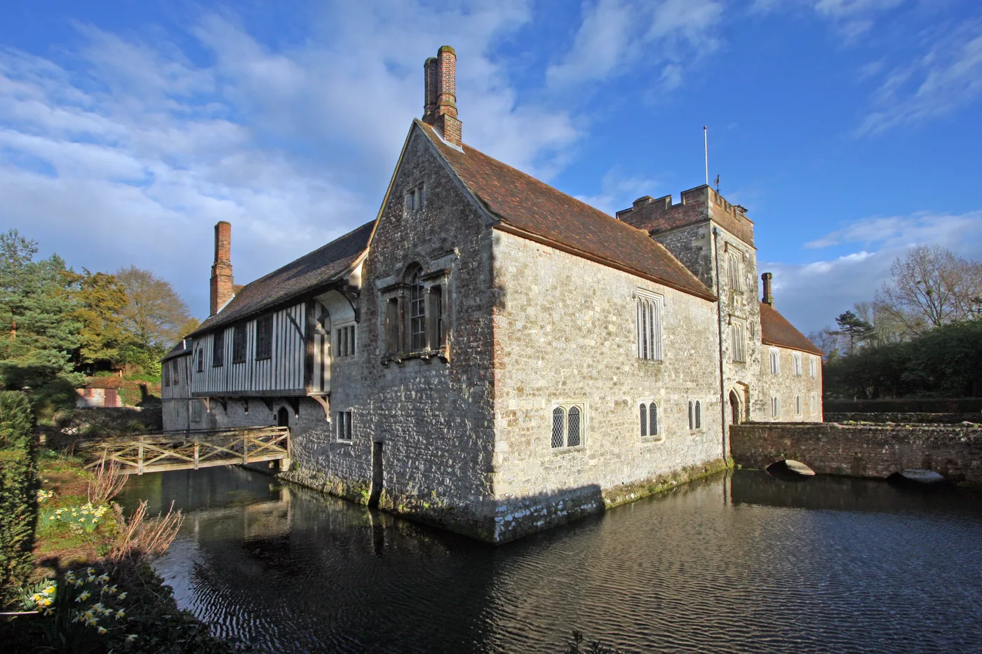 Ightham Mote across its moat, with the timber-framed upper floor and the battlemented tower behind.