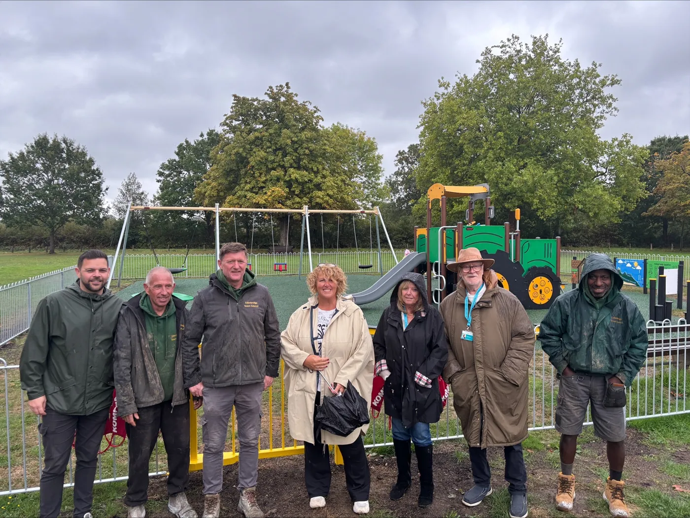 Edenbridge town councillors, council officers and grounds staff standing in front of the new Marsh Green playground on a grey day, with swings and a green tractor-shaped climbing frame behind them