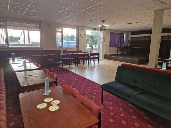 The function room at Dunton Green Social Club, with tables and chairs on a red patterned carpet, a wooden dance floor and a low stage at the far end