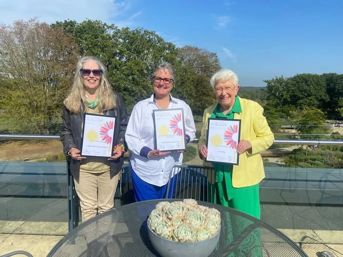 Three women standing on a terrace, each holding a framed South and South East in Bloom certificate
